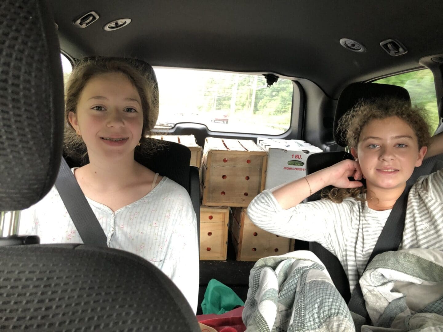 Two girls sitting in the back seat of a car, both wearing seat belts and There are cardboard boxes