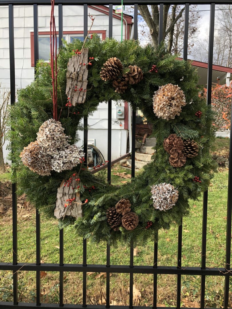 Wreath made of pine cones and other decorative items hanging on a fence in front of a house
