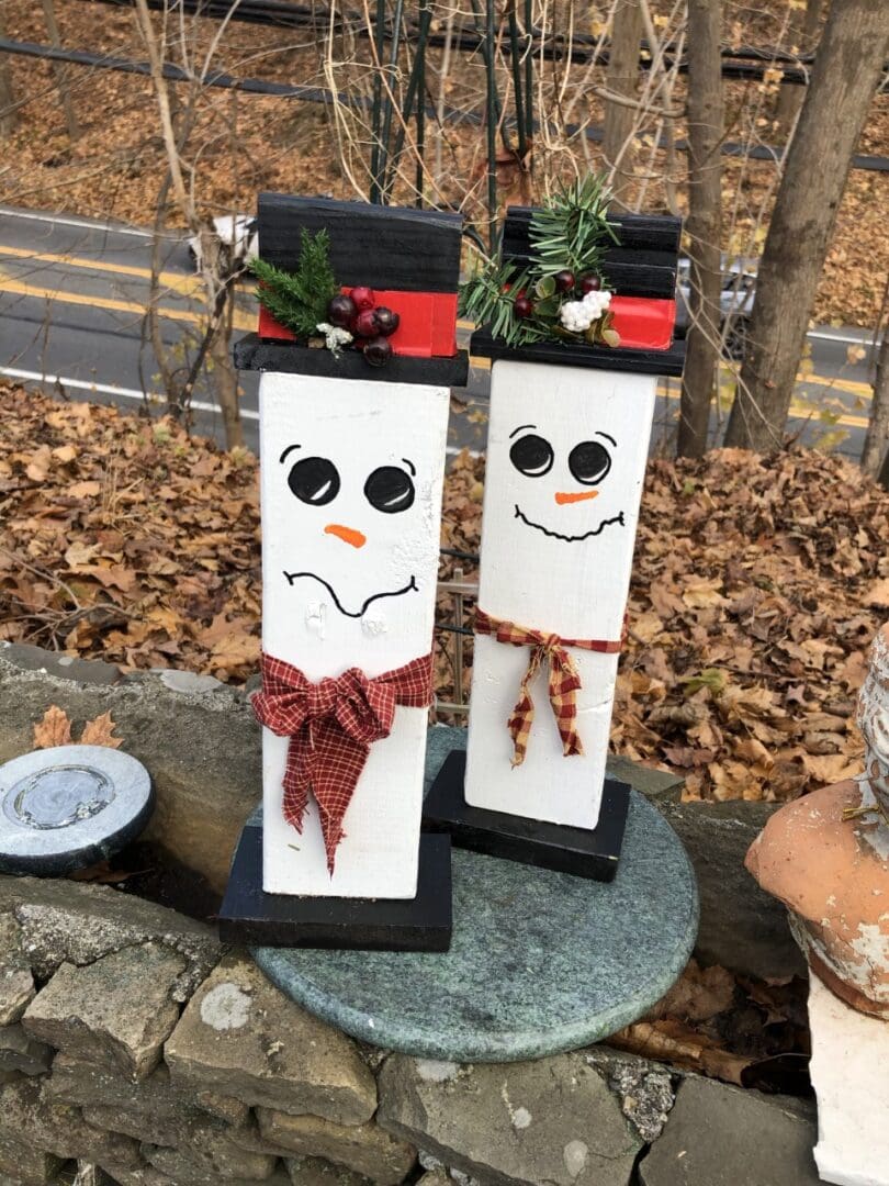 Two wooden snowmen sitting atop a stone wall, surrounded by a pot, dried leaves, a road, a fence, and trees